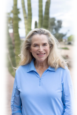 Shannon Biszantz, a Sun City Realtor with long blonde hair, smiles warmly while standing outdoors in a light blue long-sleeve shirt against a blurred backdrop of cacti and the desert landscape.